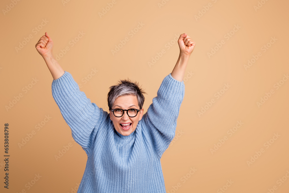 Photo portrait of attractive senior woman raise fists scream yes dressed stylish blue knitted clothes isolated on beige color background