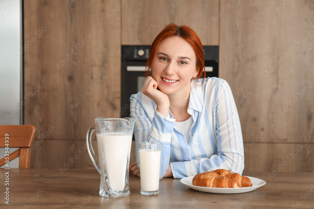 Young redhead woman with glass of milk and croissant in kitchen