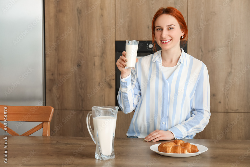 Young redhead woman with glass of milk and croissant in kitchen