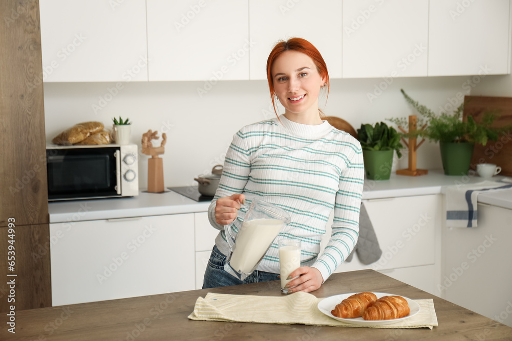 Young redhead woman pouring milk into glass in kitchen