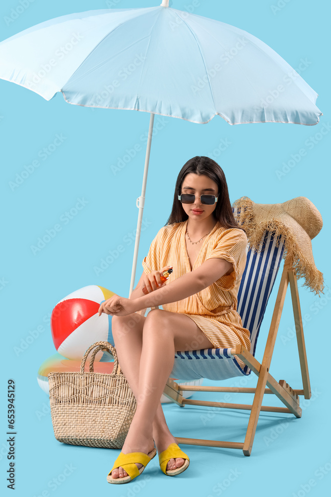 Young woman applying sunscreen cream in deck chair on blue background