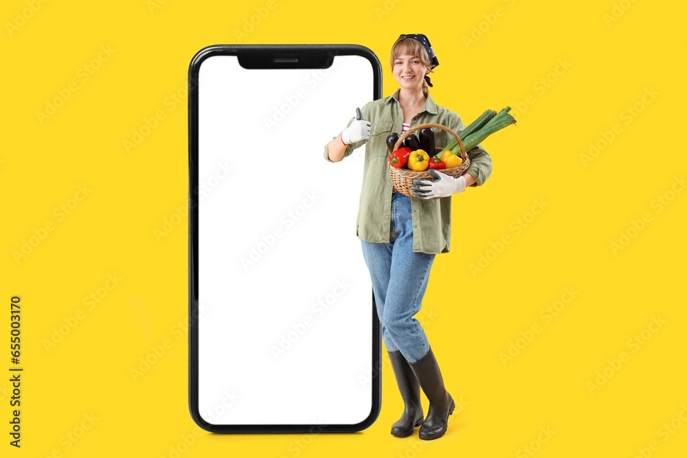 Big smartphone and female farmer with basket full of ripe vegetables on yellow background