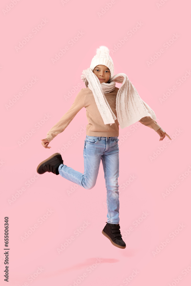 Jumping African-American boy in warm winter clothes on pink background