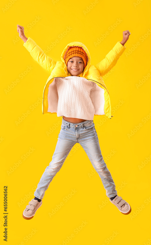 Jumping African-American boy in warm winter clothes on yellow background