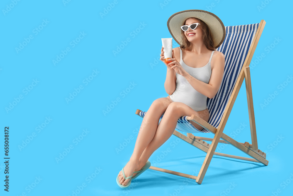 Beautiful young woman with sunscreen cream sitting on deck chair against blue background