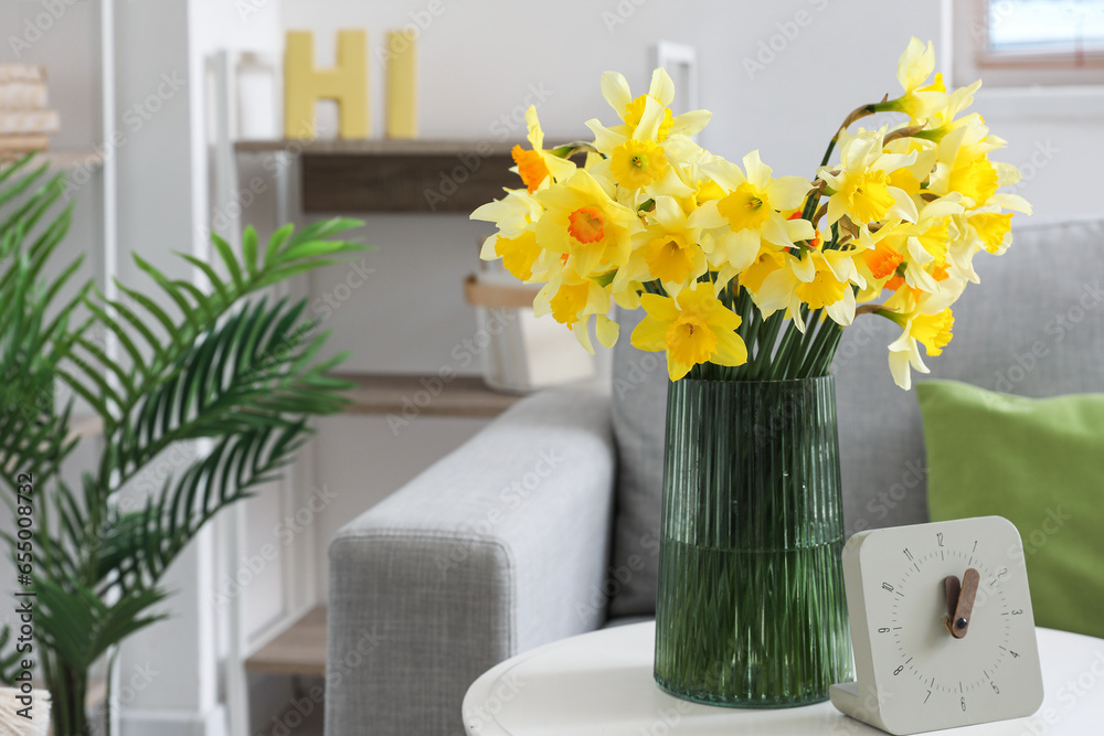 Vase with blooming narcissus flowers and clock on coffee table in interior of living room