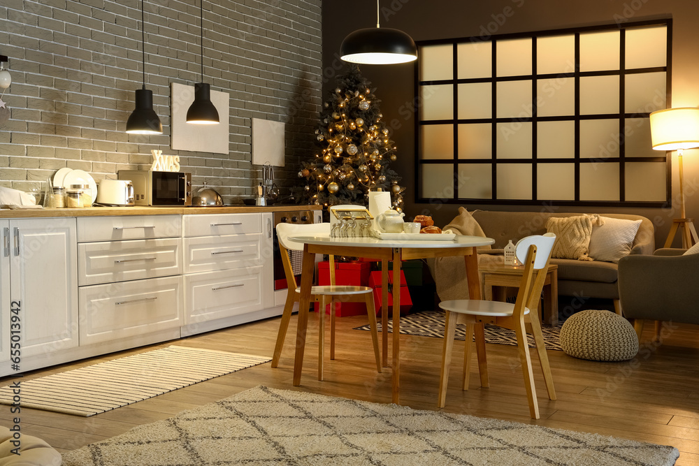 Interior of modern kitchen with Christmas tree, glowing lamps and dining table at evening