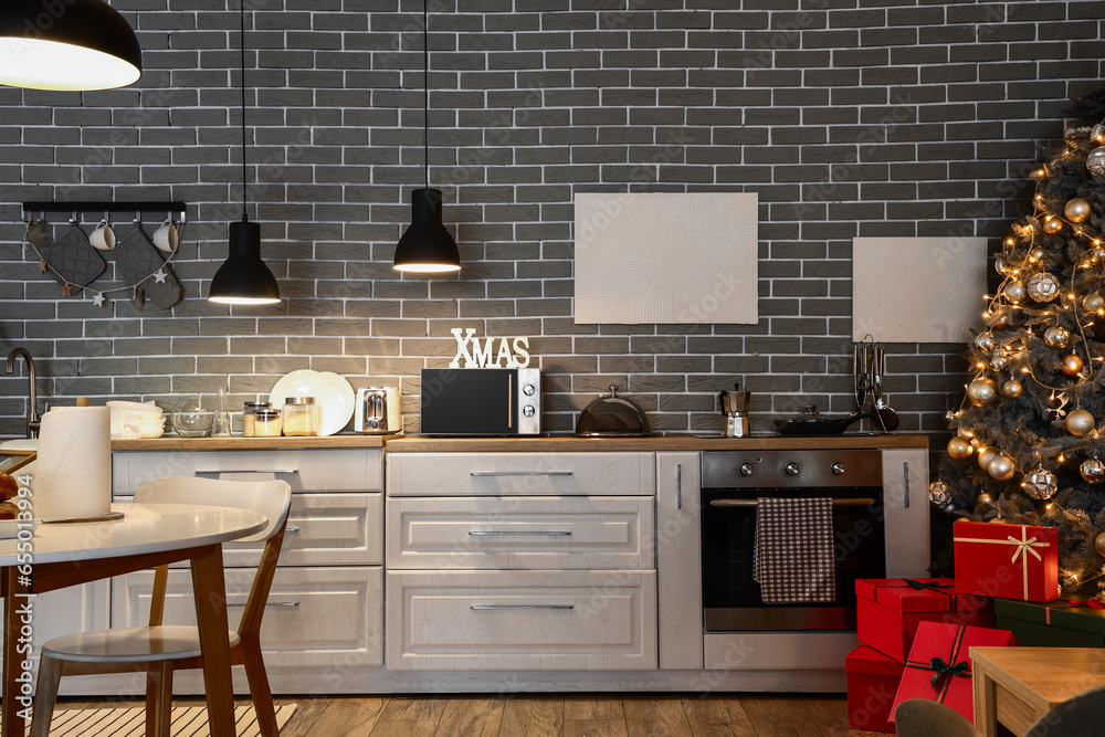 Interior of modern kitchen with Christmas tree, glowing lamps, gift boxes and white counters at evening