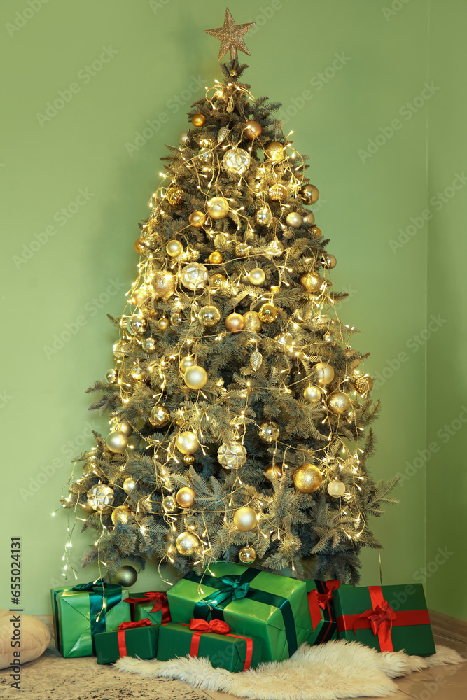 Decorated Christmas tree with gift boxes and glowing lights near green wall