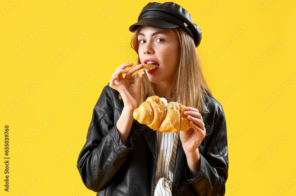 Beautiful young woman eating tasty croissant on yellow background