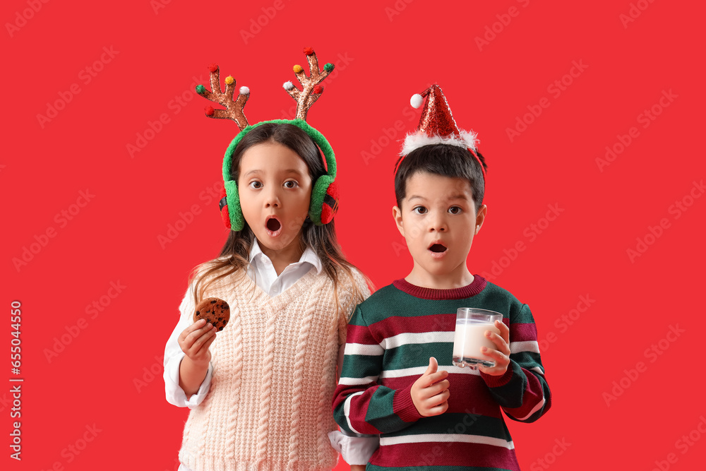 Surprised little children with Christmas decor, glass of milk and cookie on red background