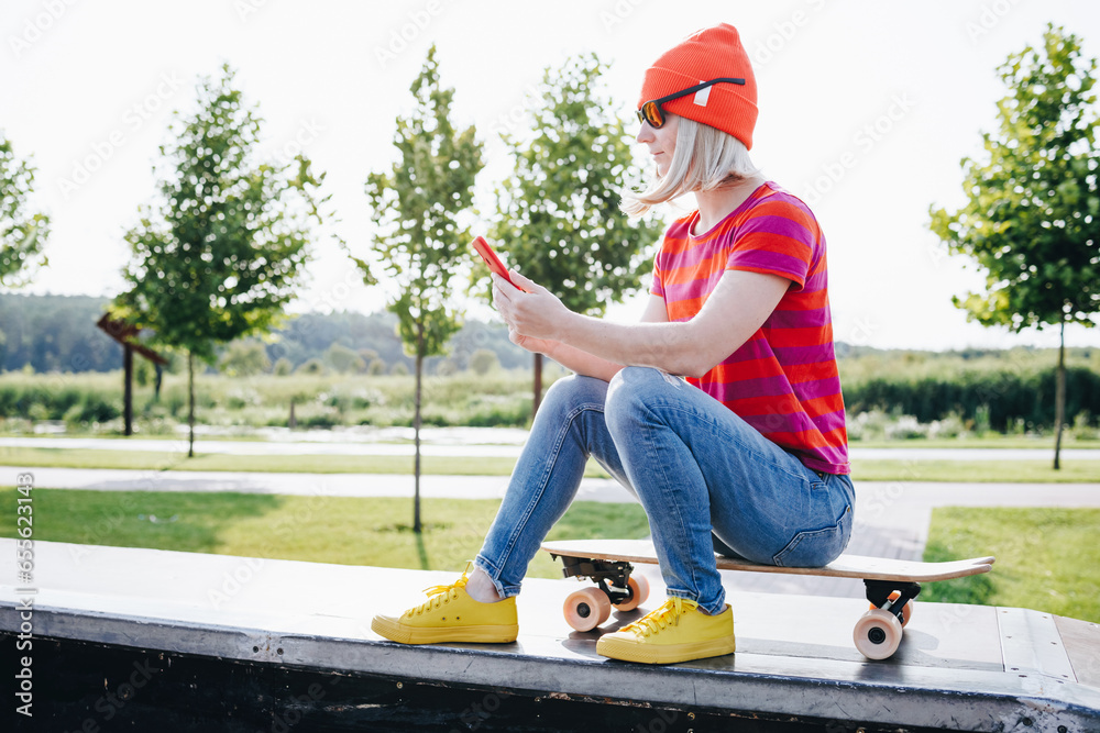 Young urban girl with her skateboard looking at smartphone outdoors. Happy young hipster woman with skateboard using mobile phone