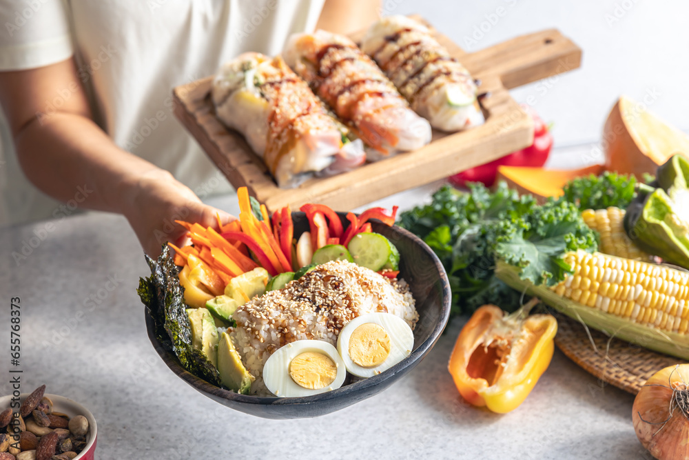 Bowl with vegetables and rice and spring rolls in female hands in the kitchen.