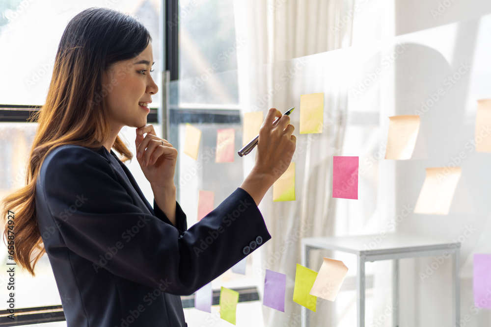 Asian businesswoman scheduling work strategy, planning work in the office with paper stickers on the glass.