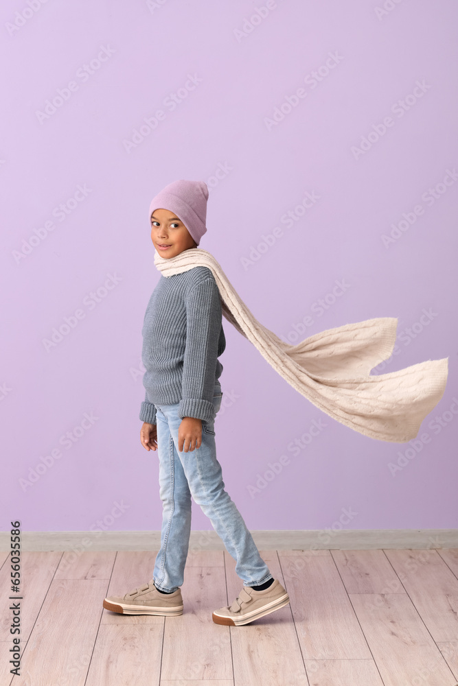 Cute African-American boy in warm winter clothes on lilac background