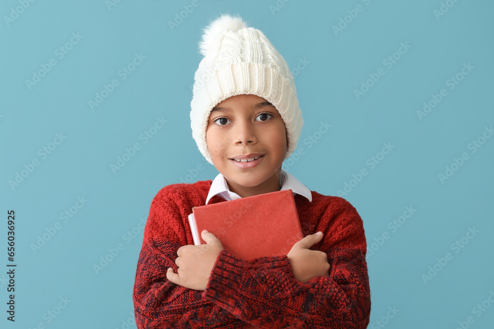 Cute African-American boy in winter clothes and with book on blue background