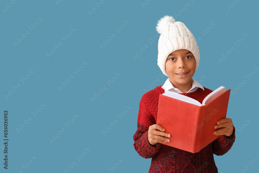 Cute African-American boy in winter clothes reading book on blue background