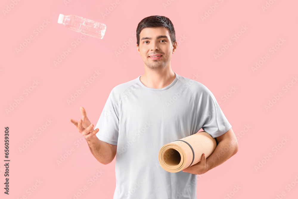 Sporty young man with bottle of water and fitness mat on pink background