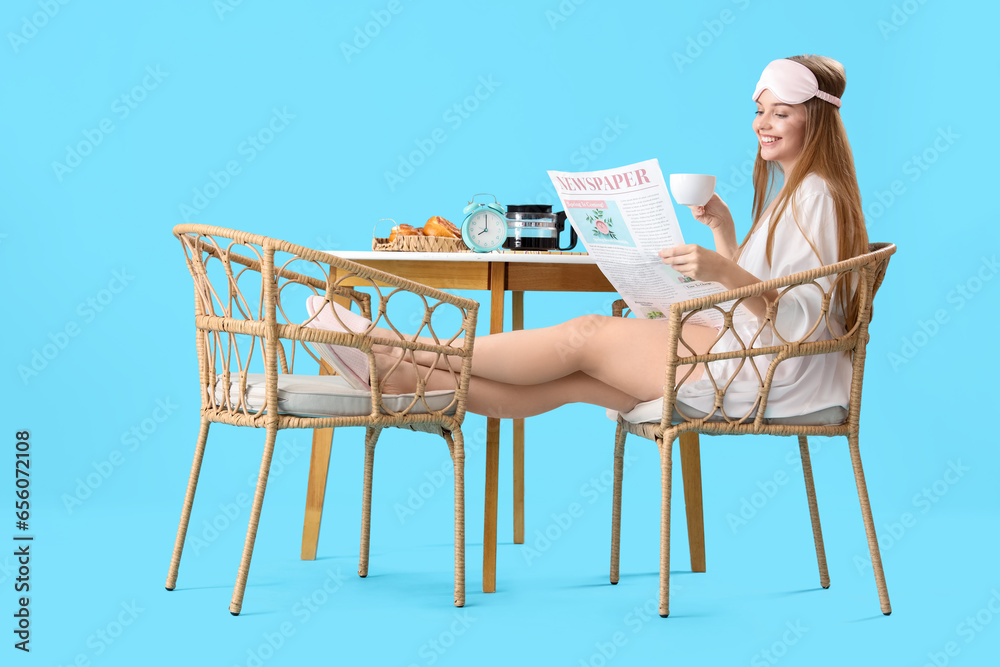 Young woman reading newspaper and drinking coffee while having breakfast on blue background