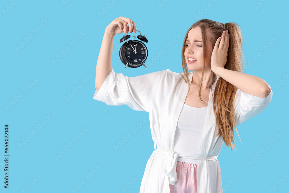 Young woman suffering from sound of loud alarm clock on blue background