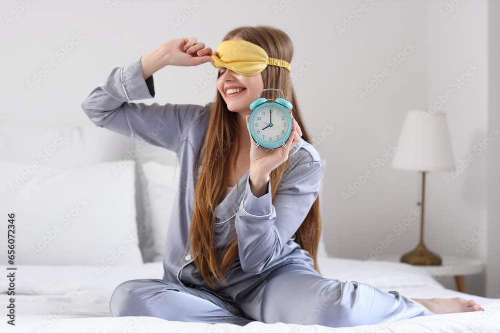 Happy young woman in pajamas with alarm clock on bed at home