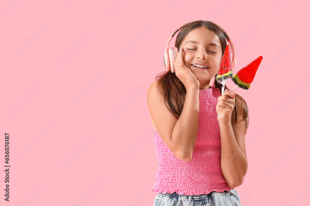 Happy little girl with lollipops in shape of watermelon slices listening music on pink background