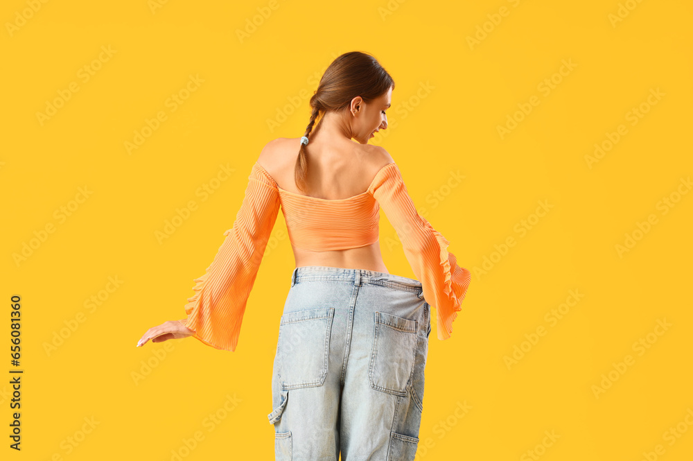 Beautiful young woman in loose jeans on yellow background, back view. Weight loss concept