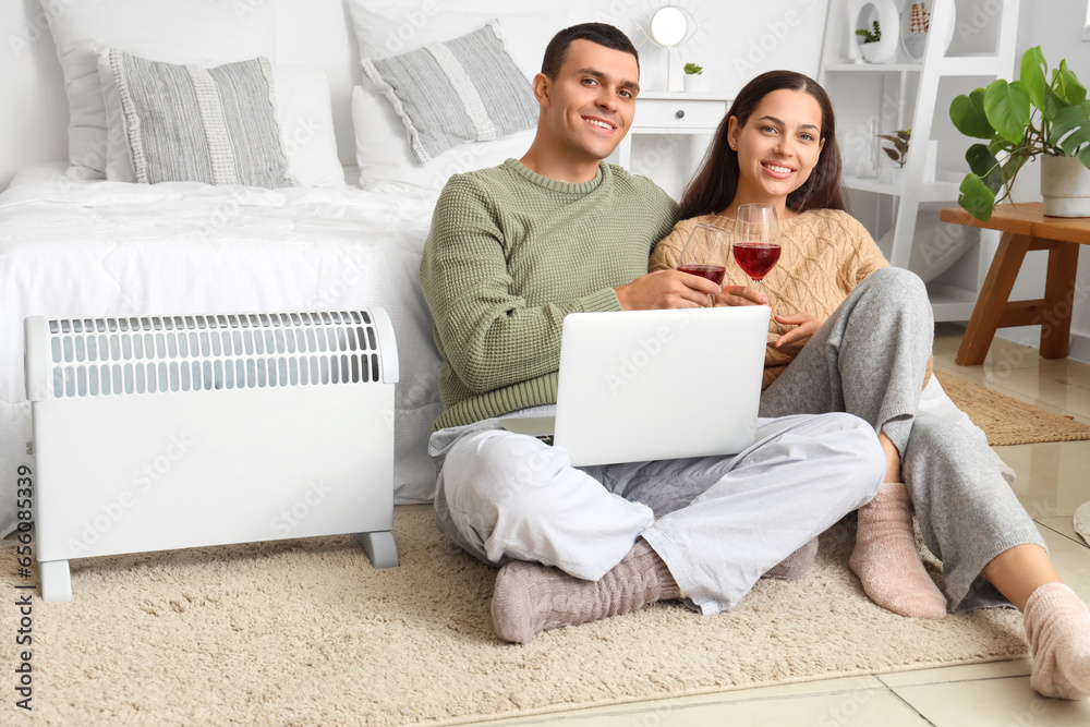 Young couple with wine and laptop warming near radiator in bedroom