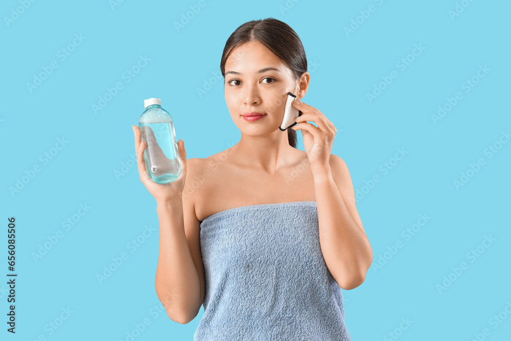 Young Asian woman with reusable cotton pad and bottle of micellar water on blue background