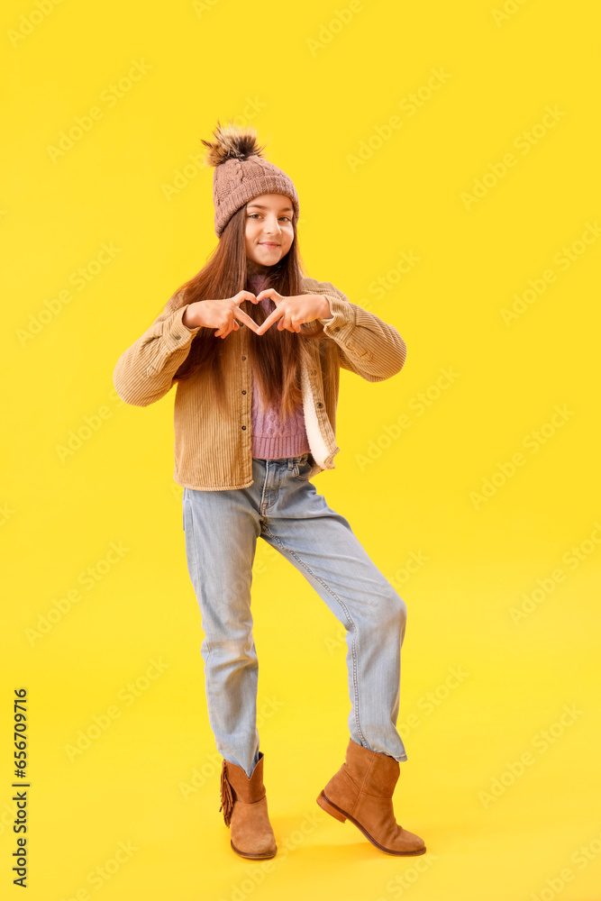 Cute little girl in winter clothes making heart shape with hands on yellow background