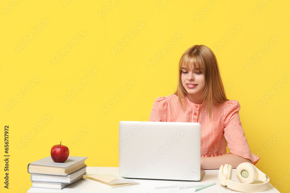 Female student with laptop doing homework at table on yellow background