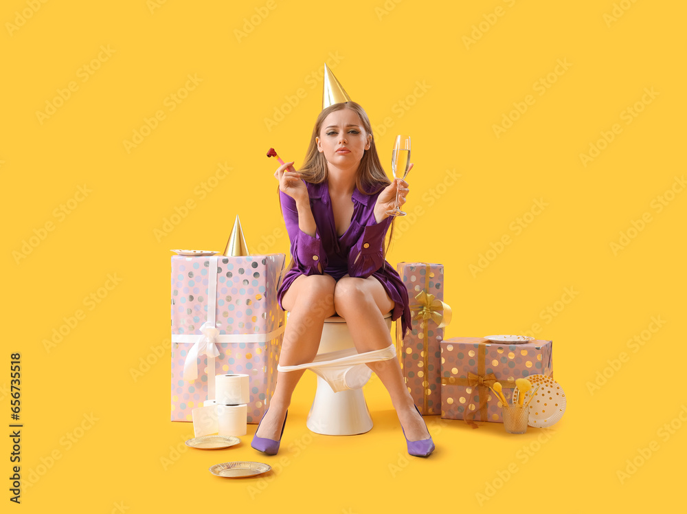 Upset young woman with champagne and party blower sitting on toilet bowl against yellow background