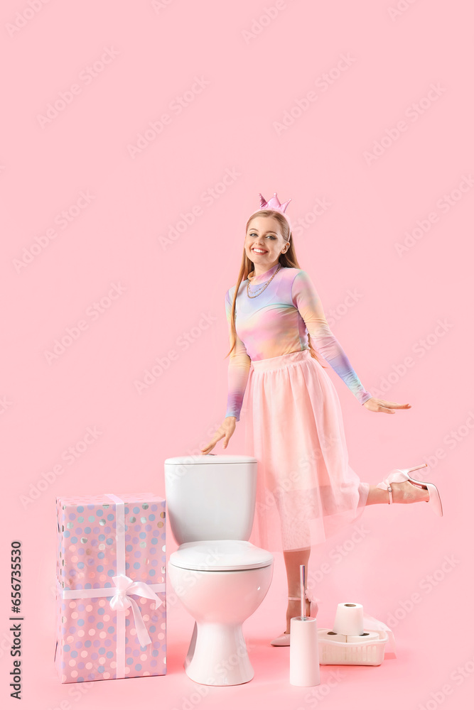 Young woman with toilet bowl and birthday gift against pink background