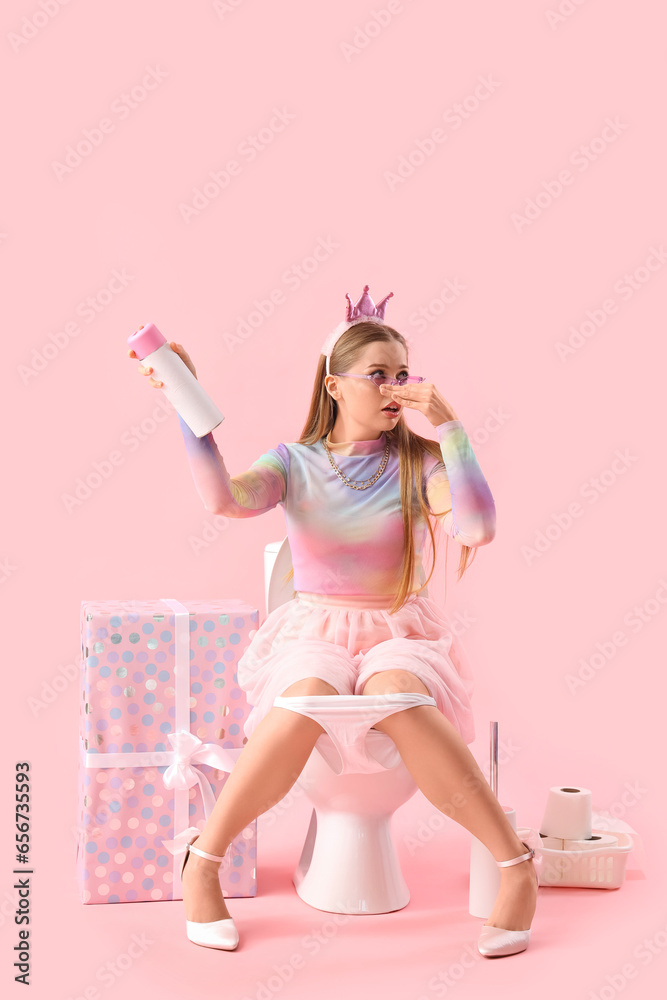 Displeased young woman with air freshener sitting on toilet bowl against pink background