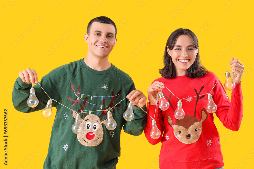 Young couple in Christmas clothes with bulb garland on yellow background