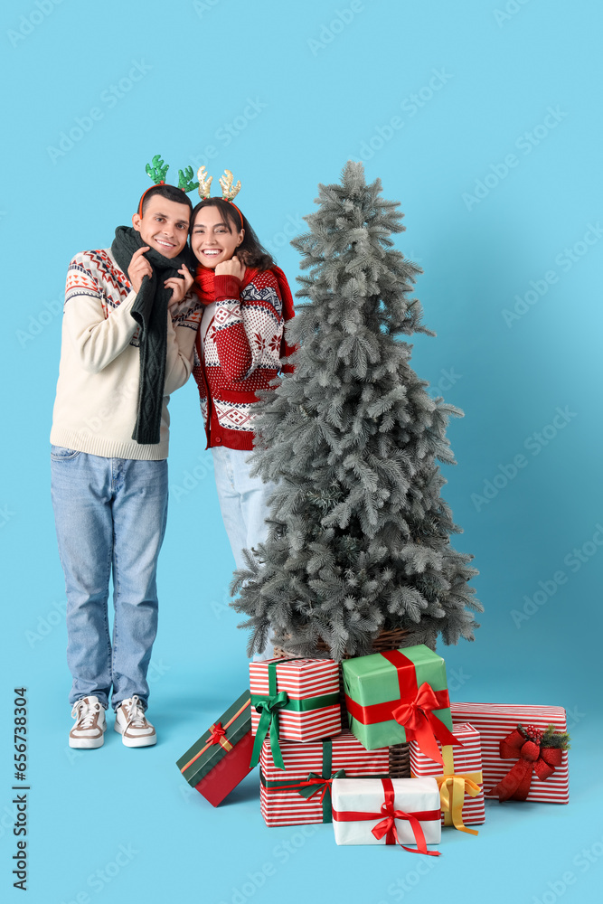 Happy young couple in reindeer horns with Christmas tree and gift boxes on blue background