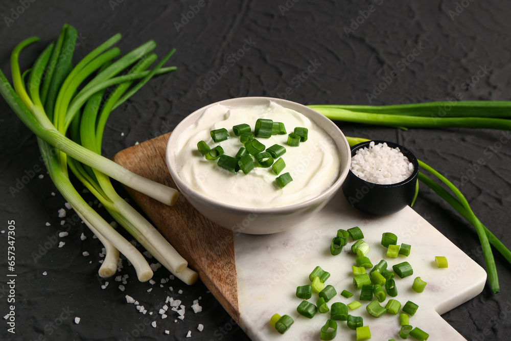 Board and bowl of tasty sour cream with sliced green onion on black background