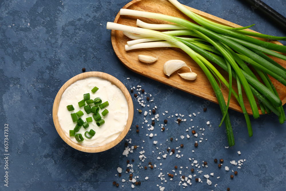 Bowl of tasty sour cream and board with green onion on blue background