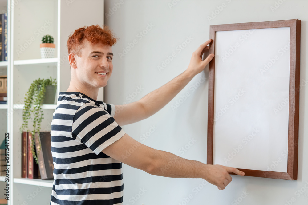 Young redhead man hanging blank frame on light wall at home