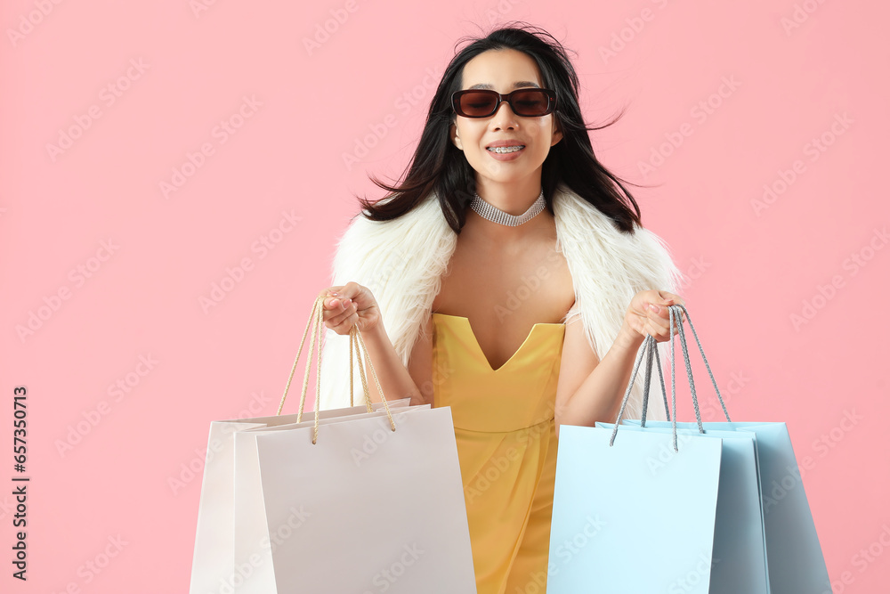 Happy beautiful young Asian woman in sunglasses with shopping bags on pink background