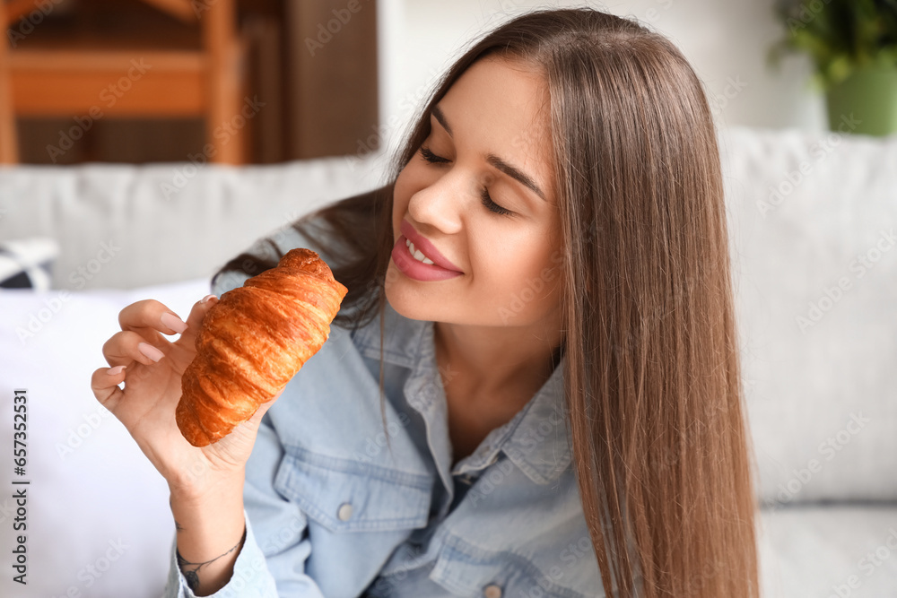 Beautiful young woman with tasty croissant sitting on sofa in living room