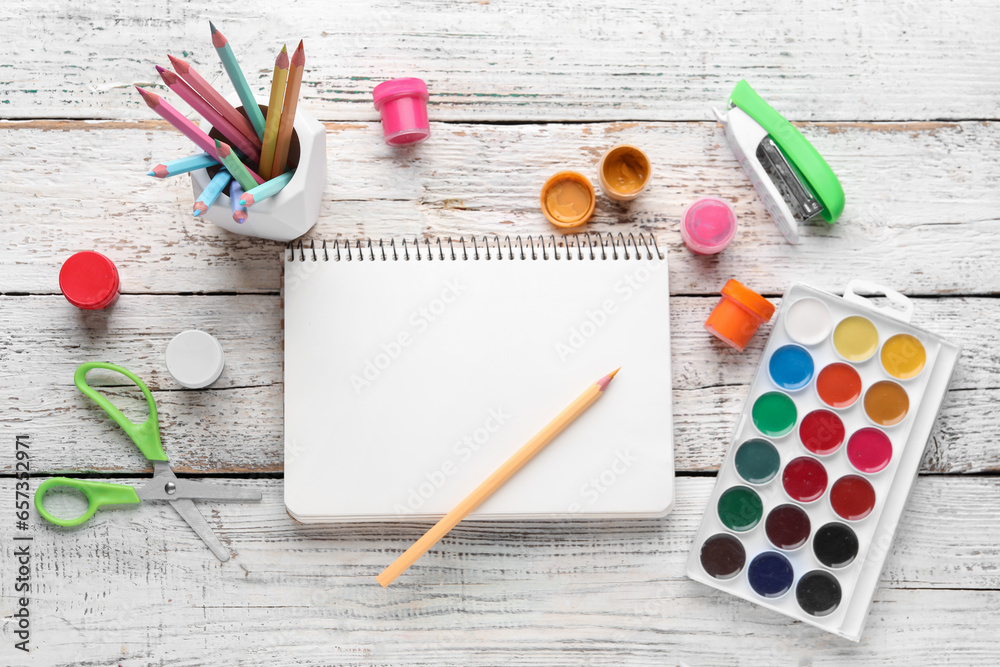 Composition with blank notebook, color pencils and paints on white wooden background