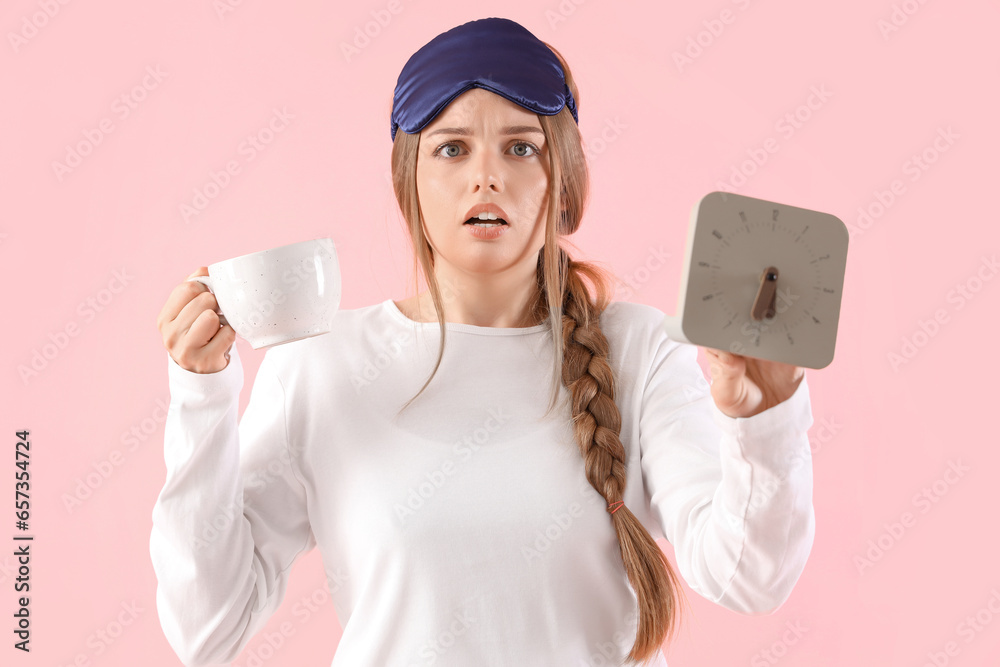 Shocked young woman in pajamas with clock and cup of coffee on pink background