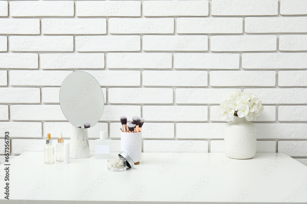 Different makeup products, mirror and beautiful flowers on dressing table near light brick wall in room