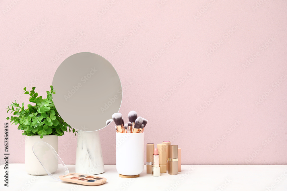 Different makeup products, mirror and houseplant on dressing table near pink wall in room