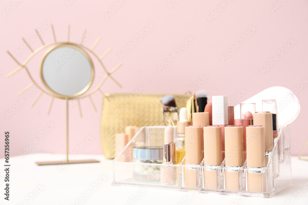 Holder with different makeup products and mirror on dressing table near pink wall in room