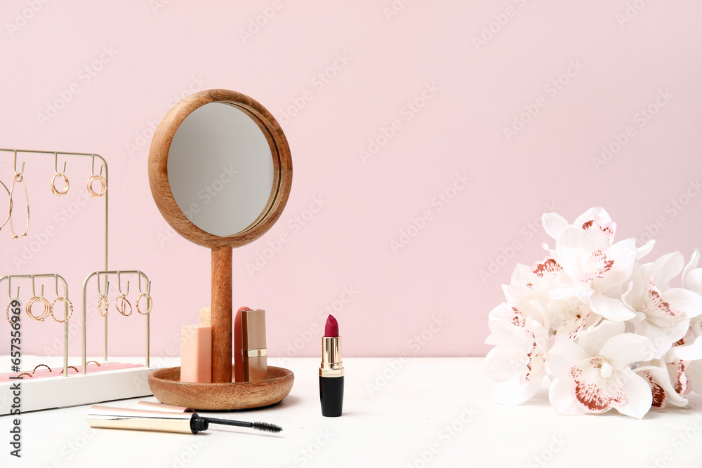 Different makeup products, mirror and beautiful flowers on dressing table near pink wall in room