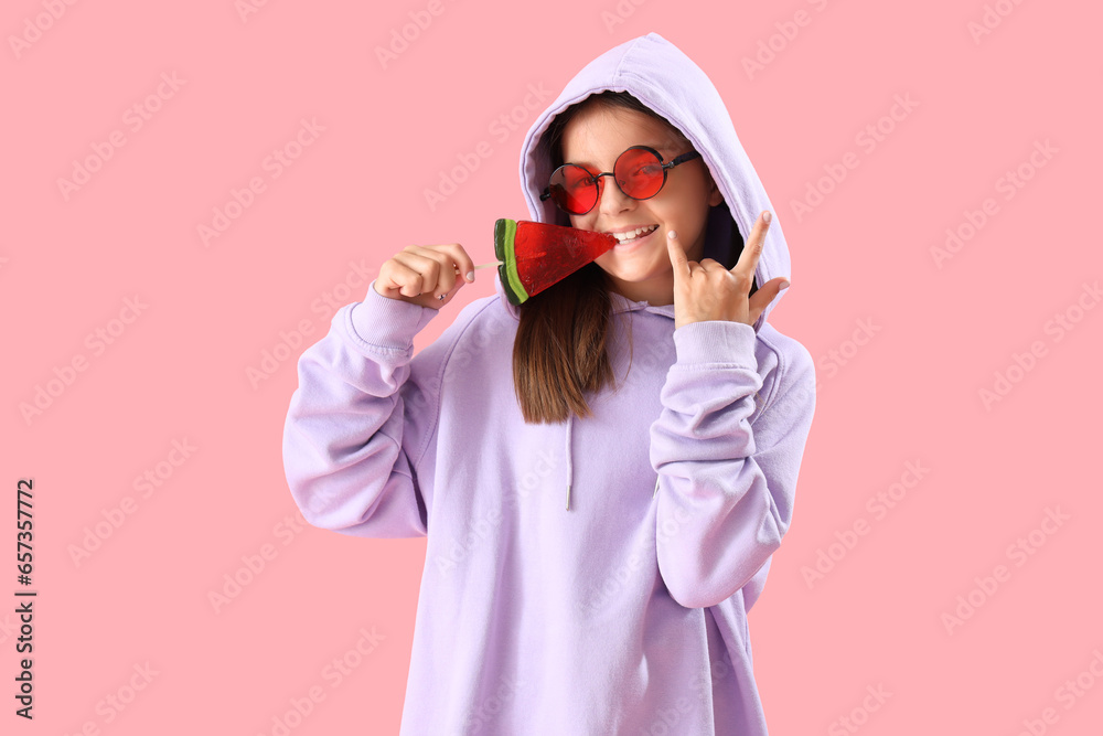 Happy little girl with lollipop in shape of watermelon slice showing  devil horns  on pink background