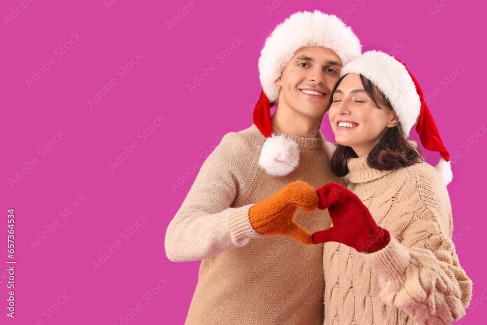 Happy young couple in Santa hats and warm gloves showing heart gesture on purple background