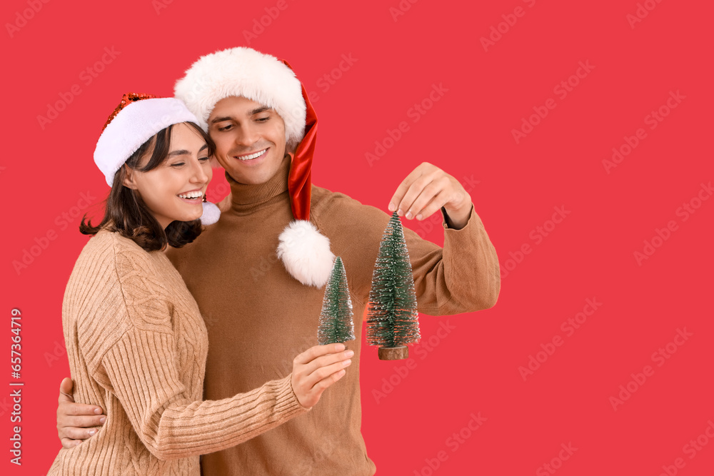 Happy young couple in Santa hats with decorative fir trees on red background